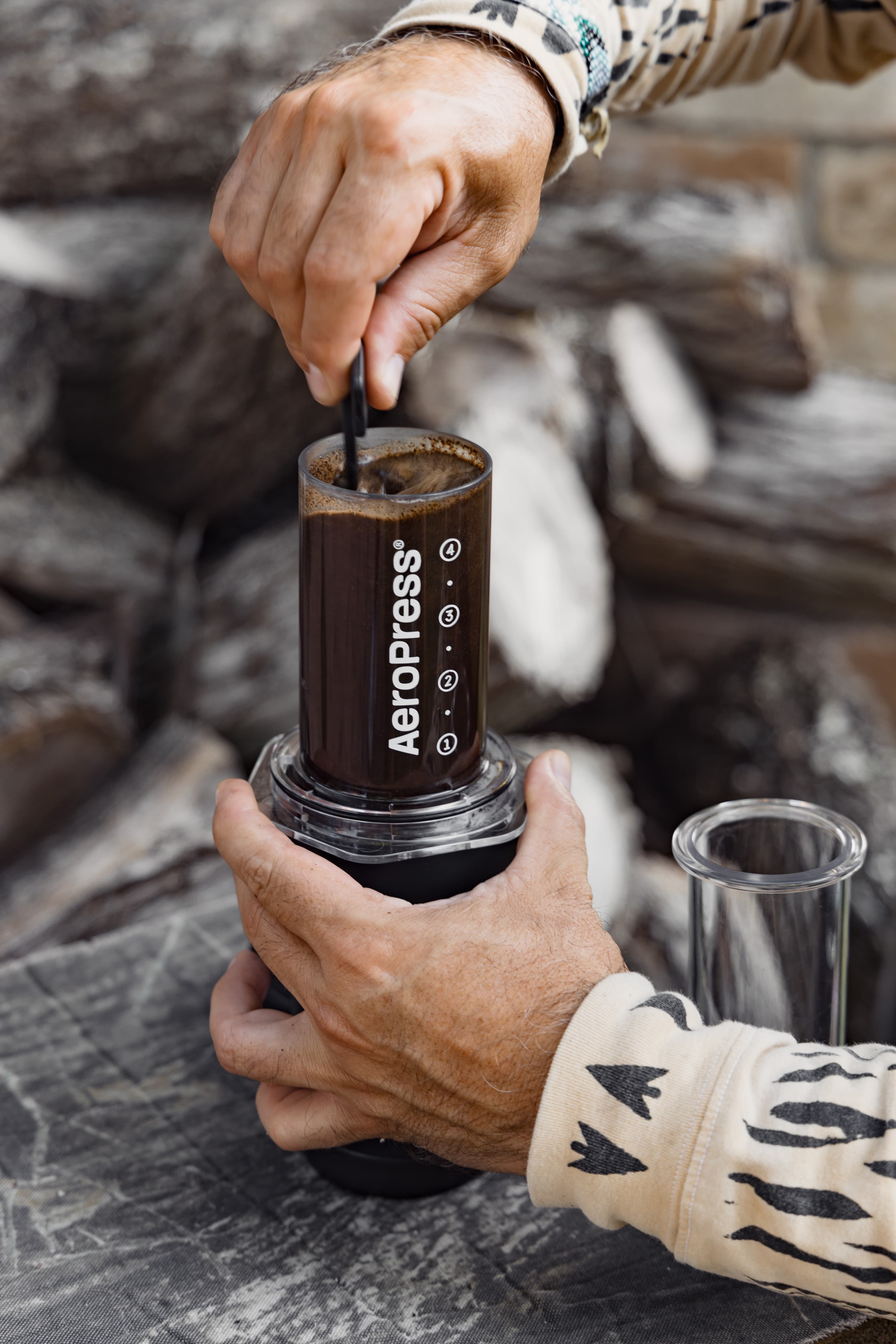 Manos revolviendo café en una AeroPress transparente en un ambiente rústico con leña apilada.