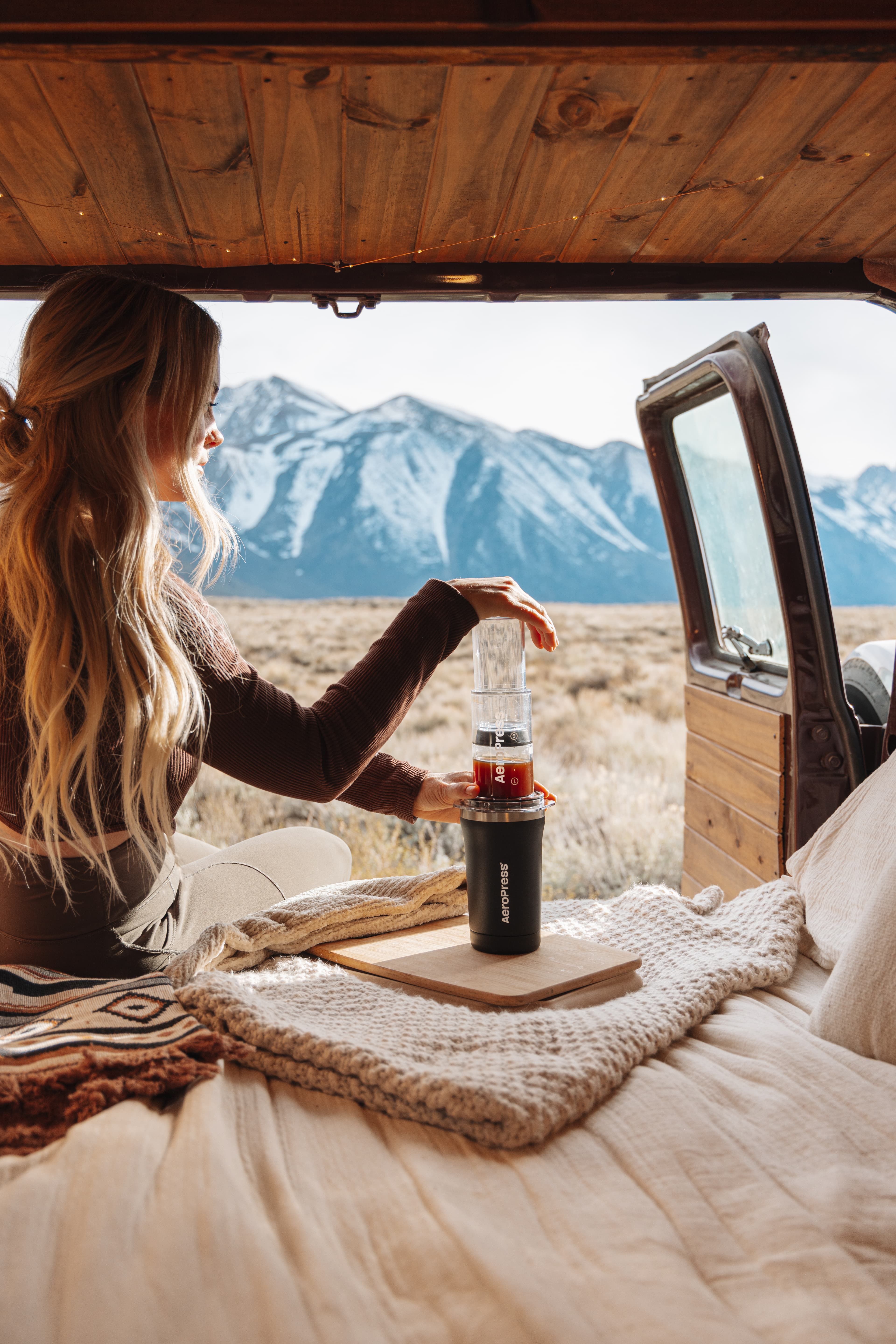 Mujer preparando café con AeroPress dentro de una furgoneta con vistas a montañas nevadas.