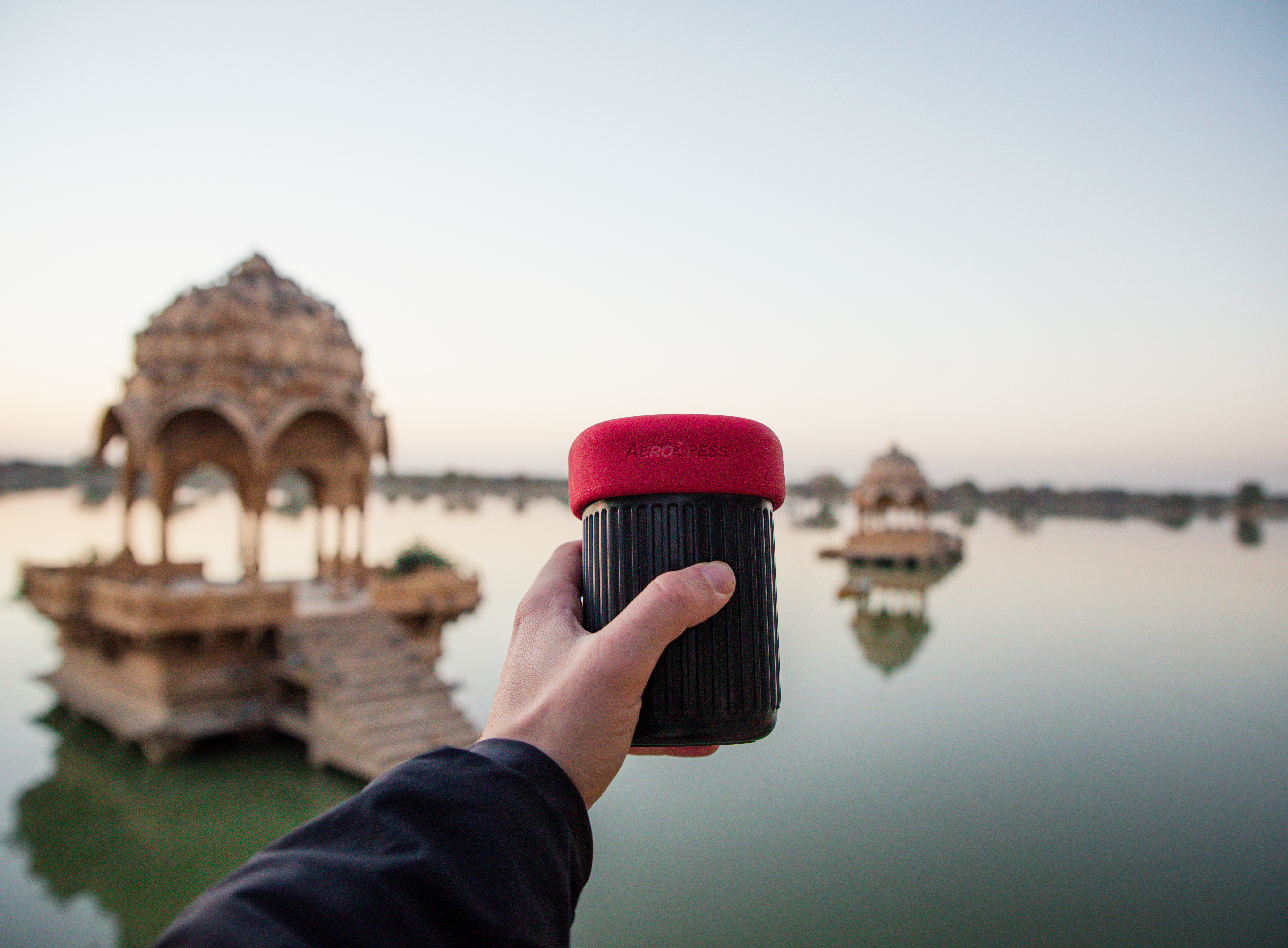 Mano sosteniendo una cafetera AeroPress frente a un lago con templos indios al atardecer.