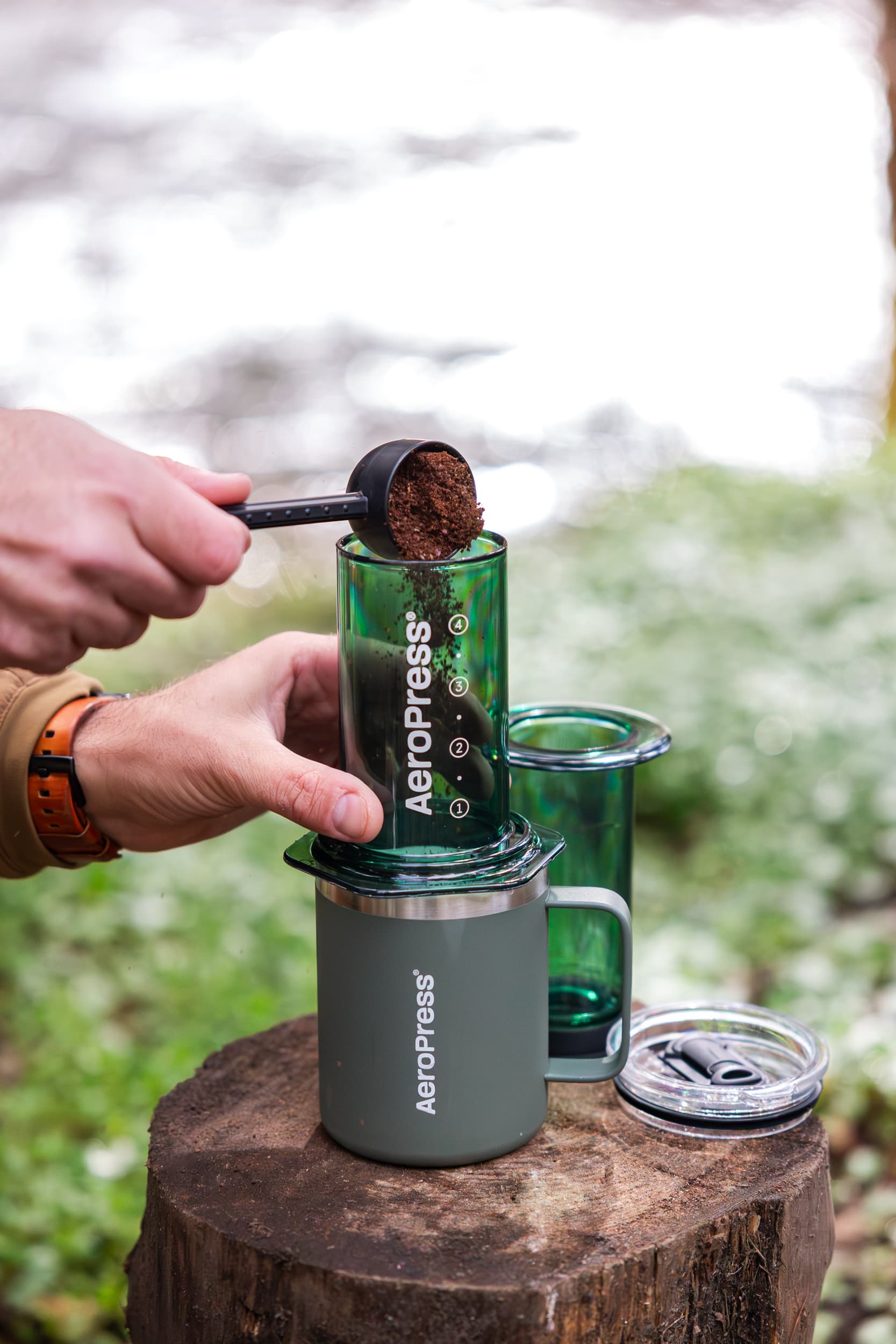 Manos vertiendo café molido en una AeroPress verde sobre una taza en un tronco.