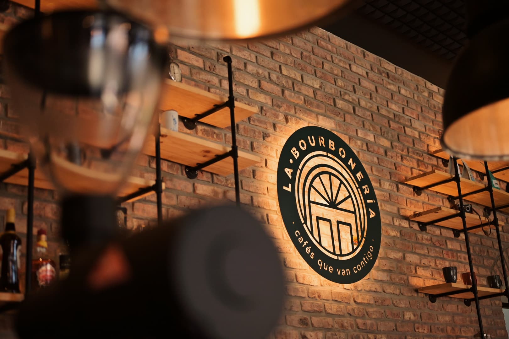 Logotipo iluminado de La Bourboneria en pared de ladrillo con estanterías de madera rústicas.