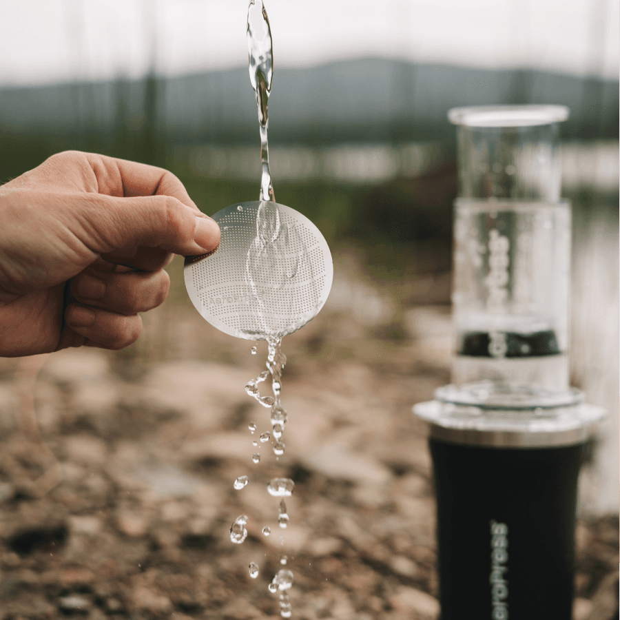 Mano enjuagando un filtro metálico AeroPress bajo un chorro de agua en el exterior.