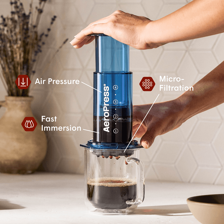 Manos presionando una cafetera AeroPress azul sobre una taza de cristal con café recién hecho.
