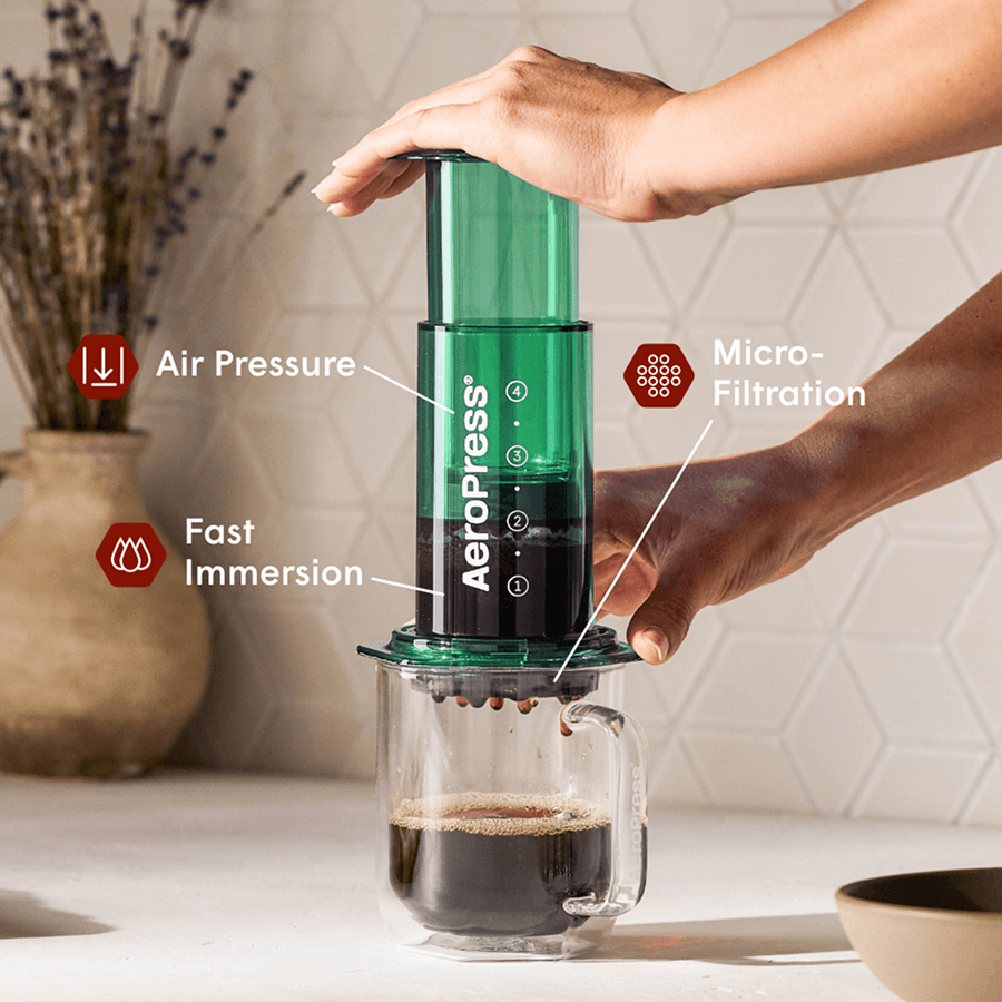 Manos presionando una cafetera AeroPress verde para filtrar café recién hecho en una taza transparente.