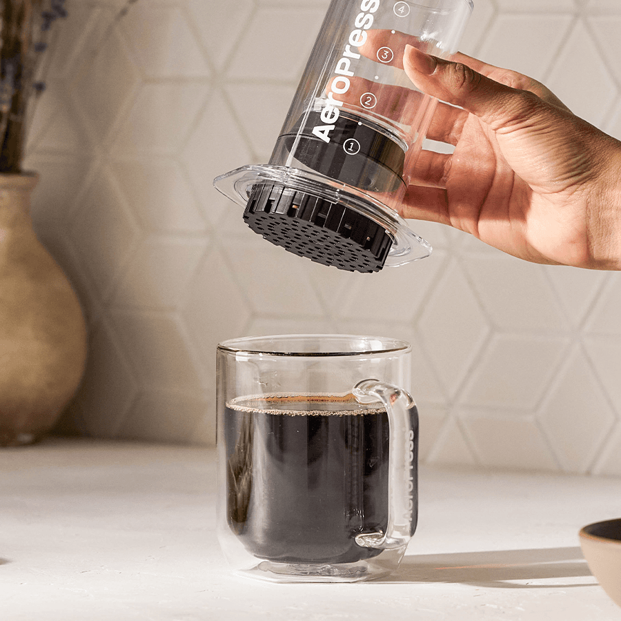 Mano sosteniendo una cafetera AeroPress sobre una taza de cristal llena de café negro.