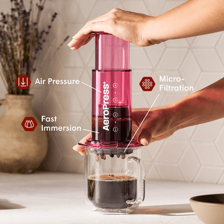 Persona presionando una cafetera AeroPress rosa para filtrar café en una taza de cristal transparente.