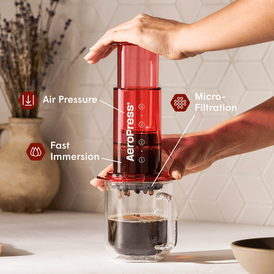 Manos presionando una cafetera AeroPress roja sobre una taza de cristal con café goteando.
