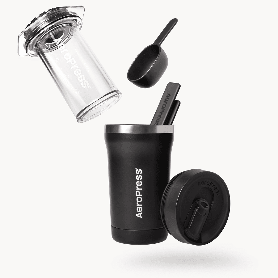 Cafetera AeroPress Go con taza de viaje negra, cuchara y agitador flotando sobre fondo blanco.