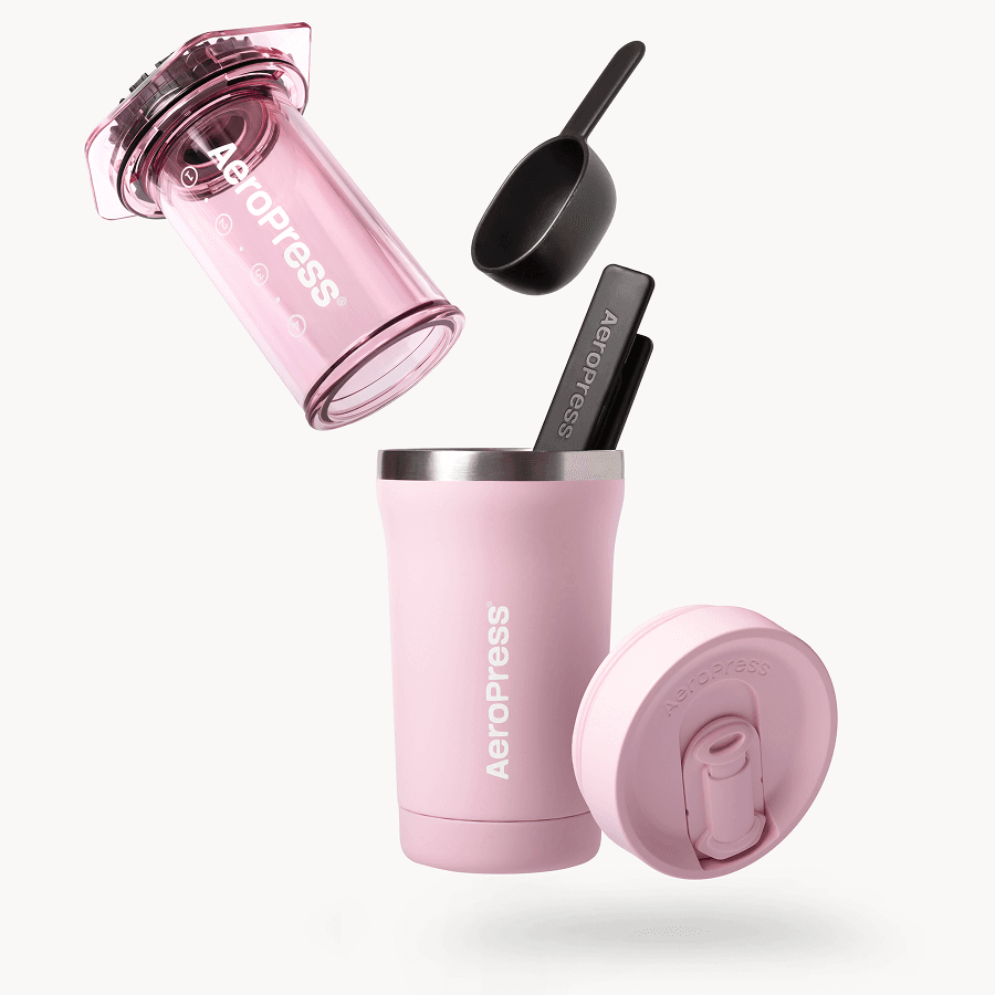 Cafetera AeroPress Go rosa con taza de viaje, tapa, cuchara y agitador sobre fondo blanco.