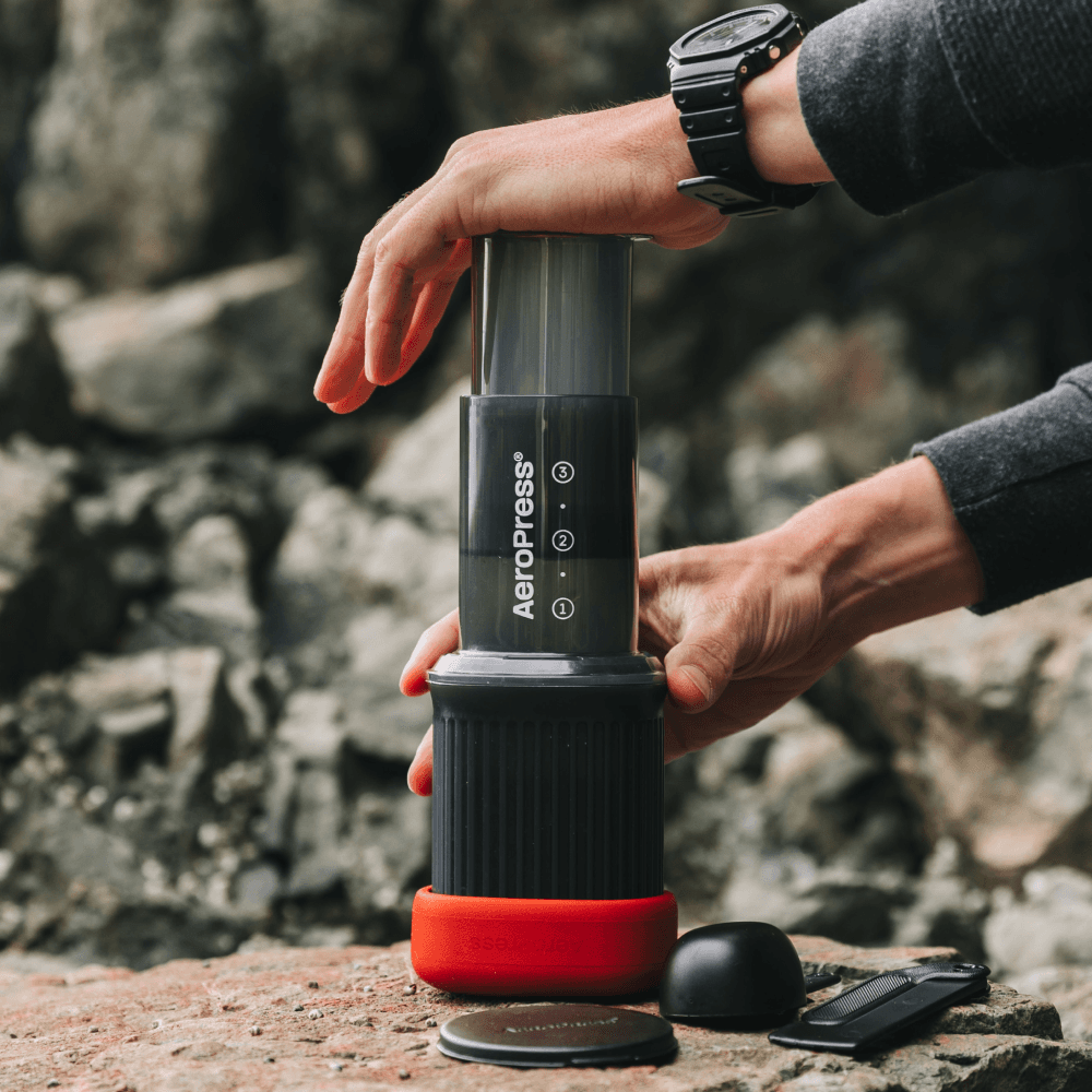 Manos presionando una cafetera portátil AeroPress sobre una roca en un paisaje exterior rocoso.