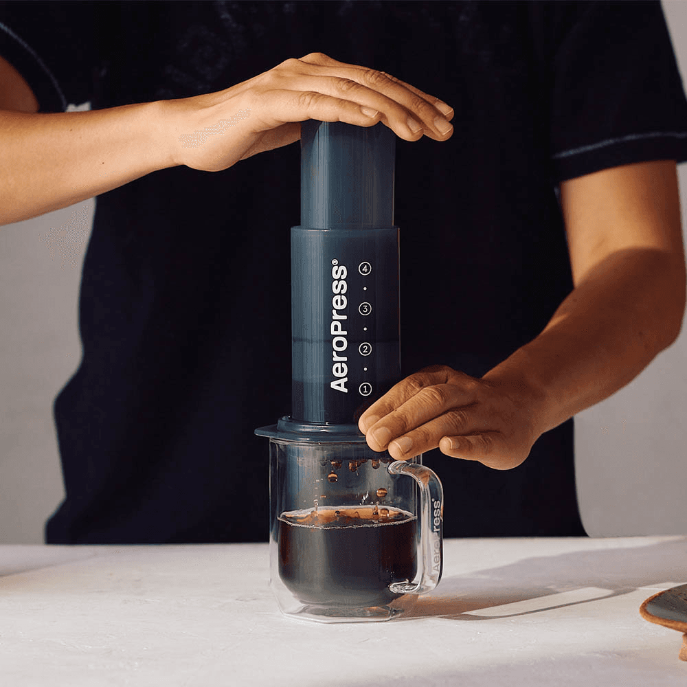 Persona presionando una cafetera AeroPress para filtrar café en una taza de cristal transparente.