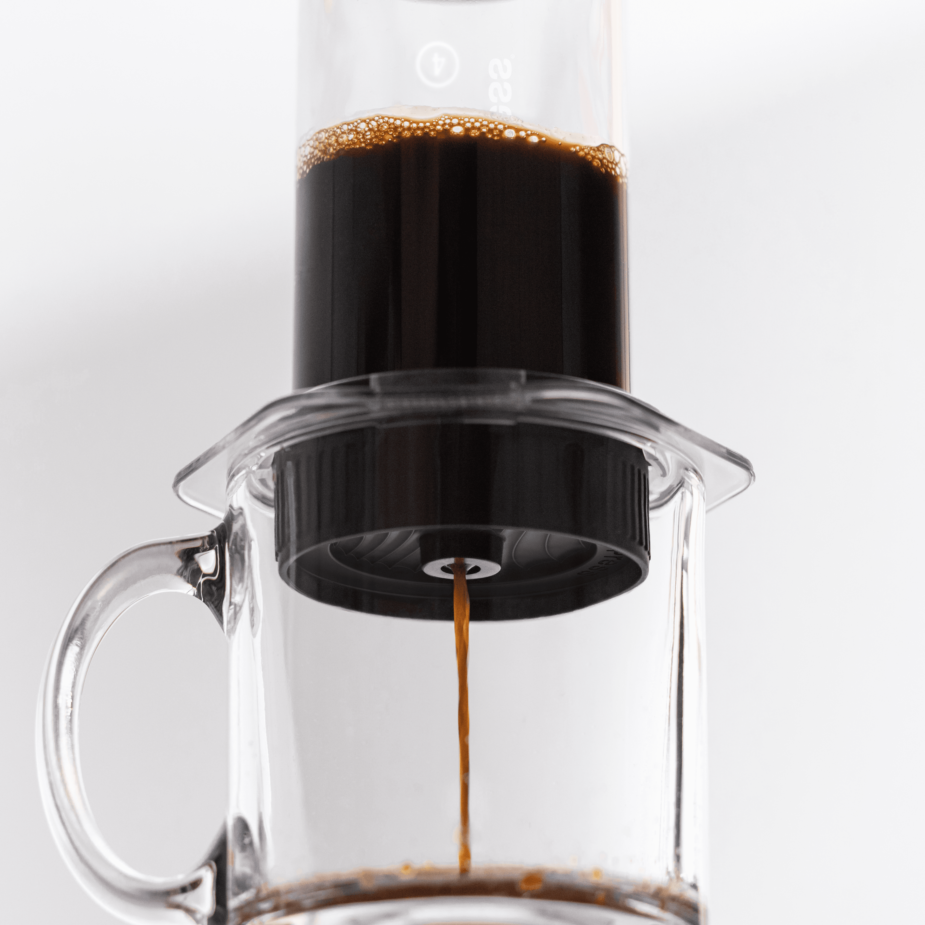 Café oscuro goteando de una cafetera AeroPress transparente hacia una taza de cristal transparente.