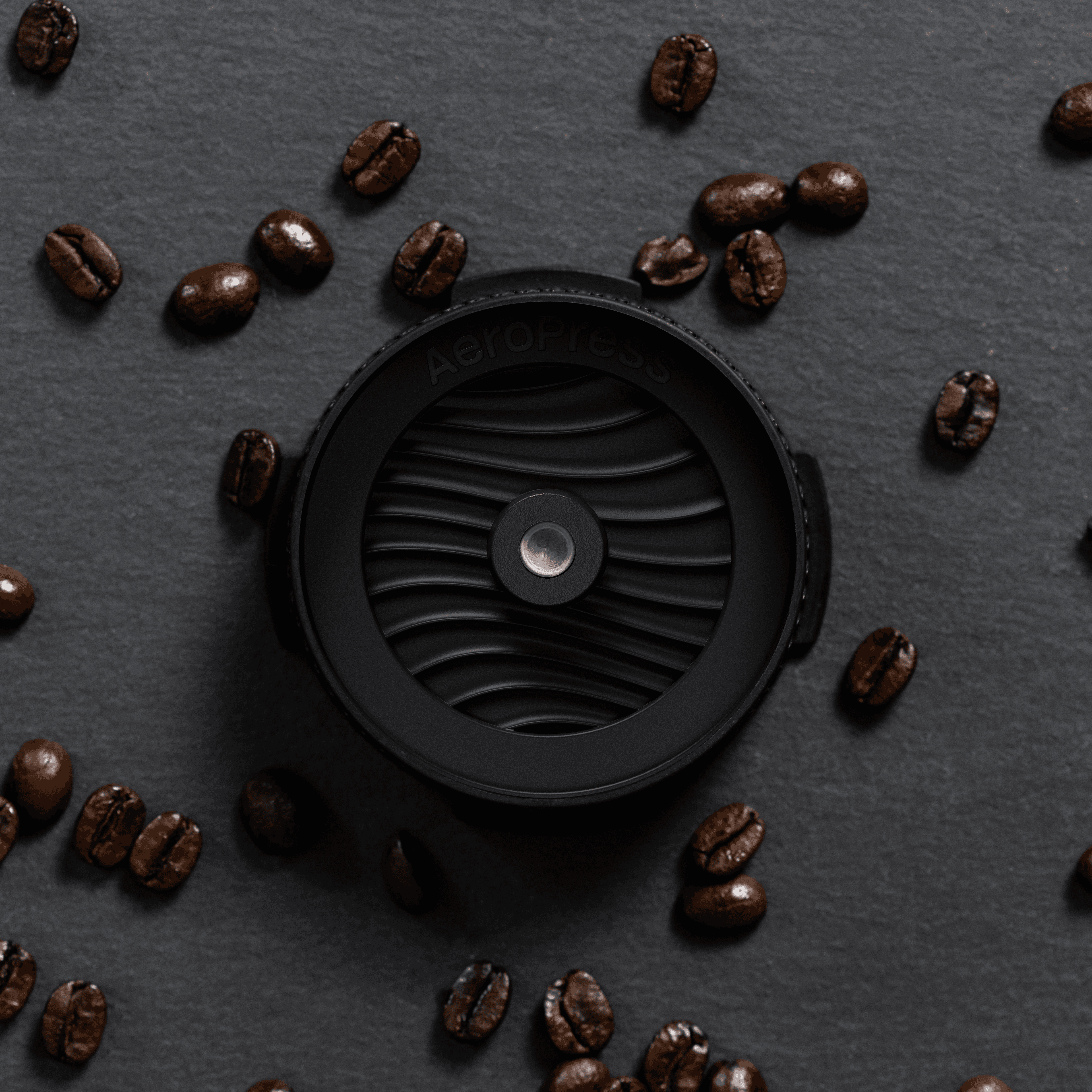 Tapa negra AeroPress con diseño ondulado rodeada de granos de café sobre una superficie oscura.