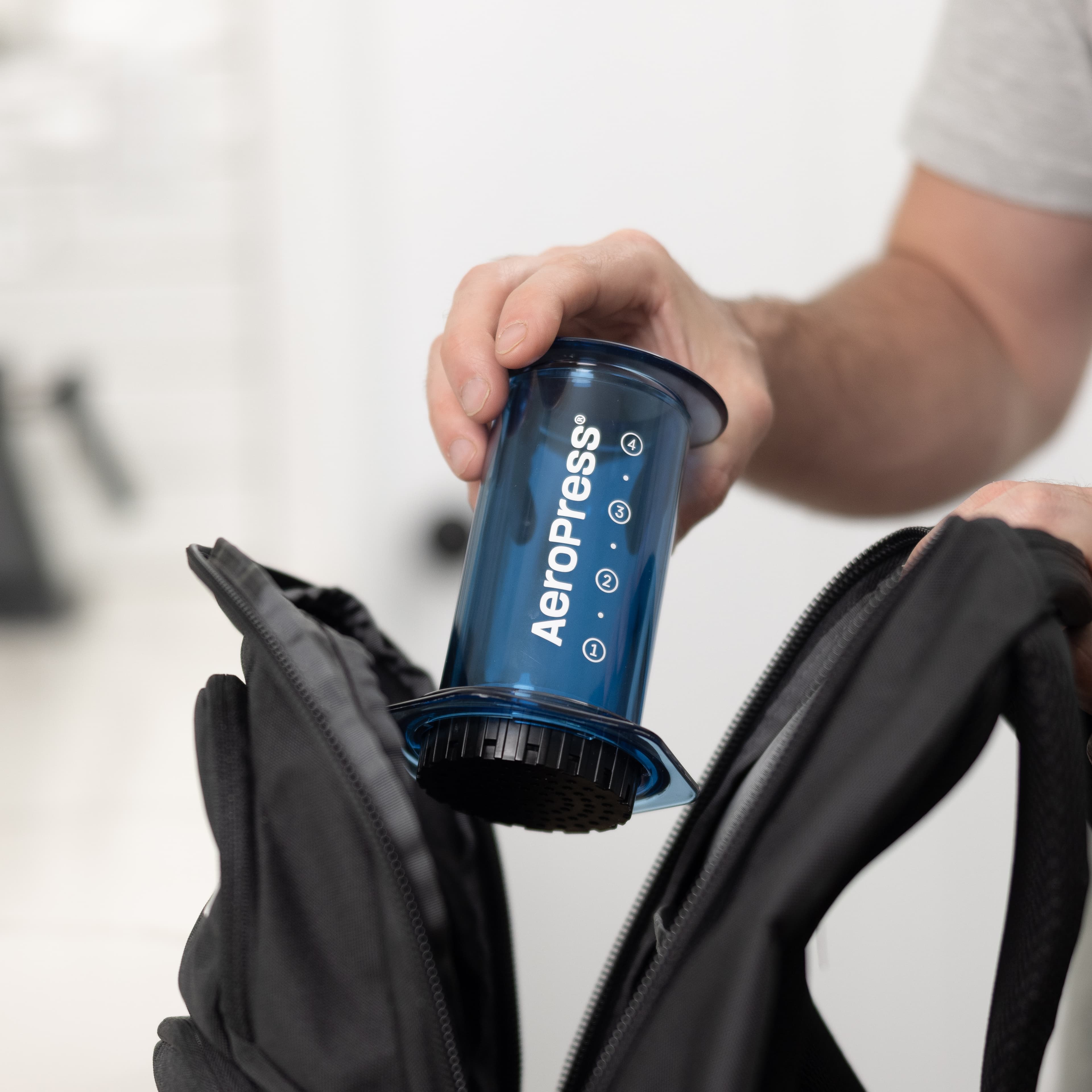 Una mano coloca una cafetera AeroPress azul transparente dentro de una mochila negra abierta.