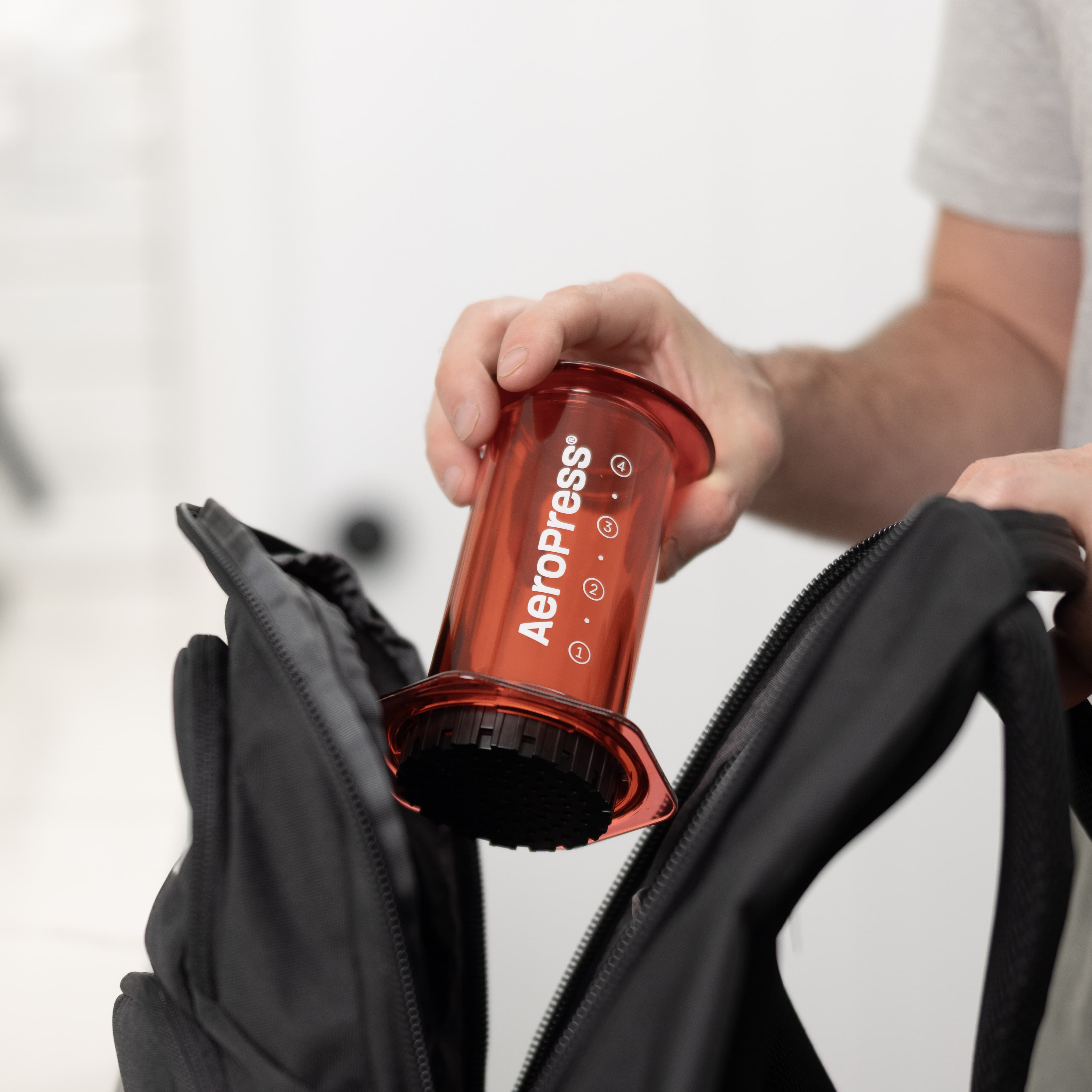 Mano colocando una cafetera AeroPress roja translúcida dentro de una mochila negra abierta.