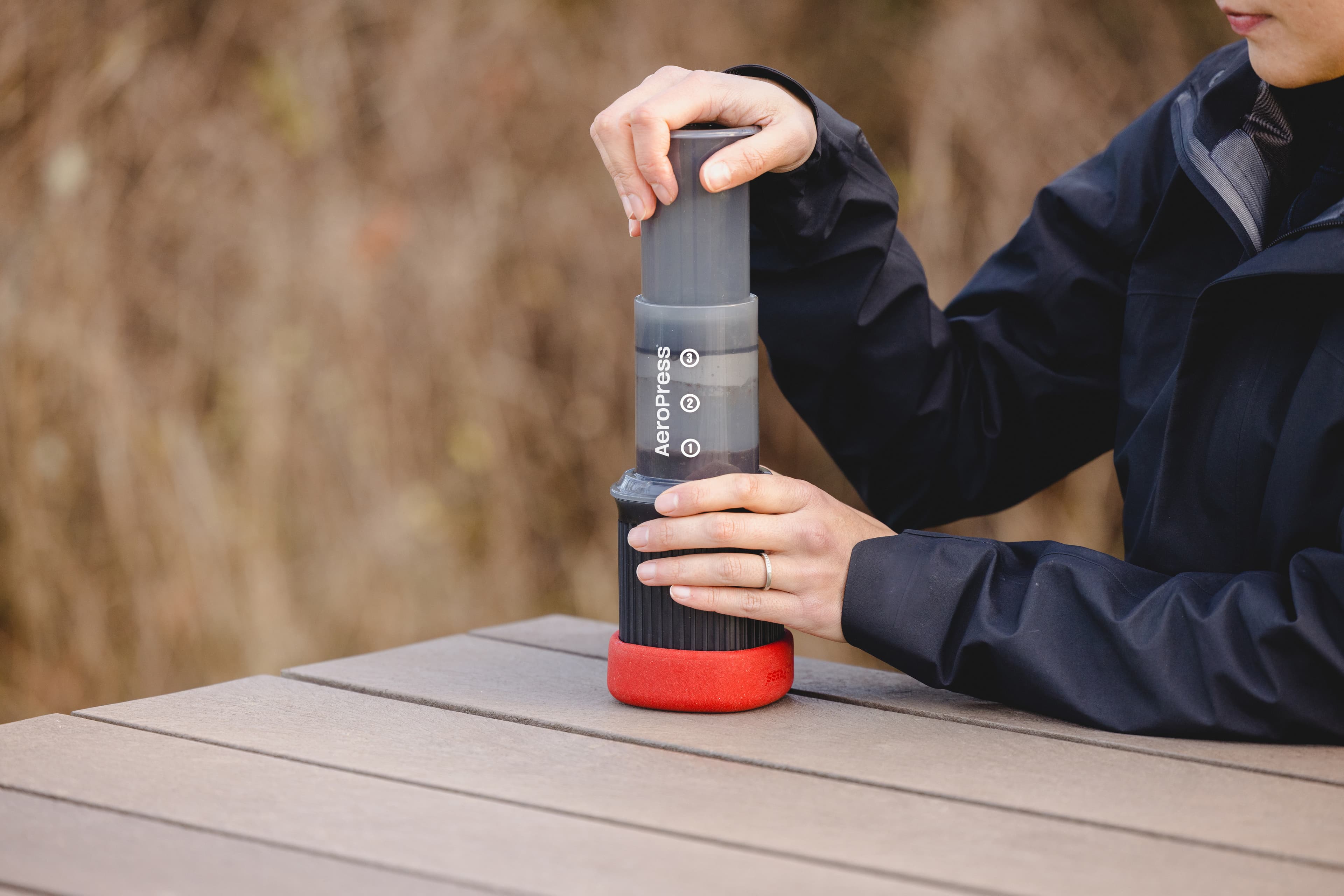Persona preparando café con una AeroPress Go sobre una mesa de madera al aire libre.