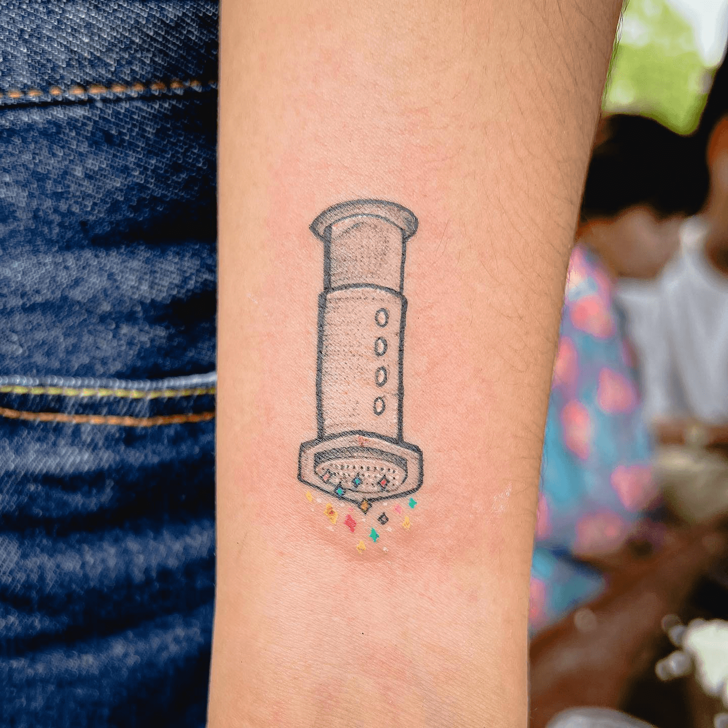 Tatuaje pequeño de una cafetera AeroPress en un antebrazo con confeti de colores cayendo.