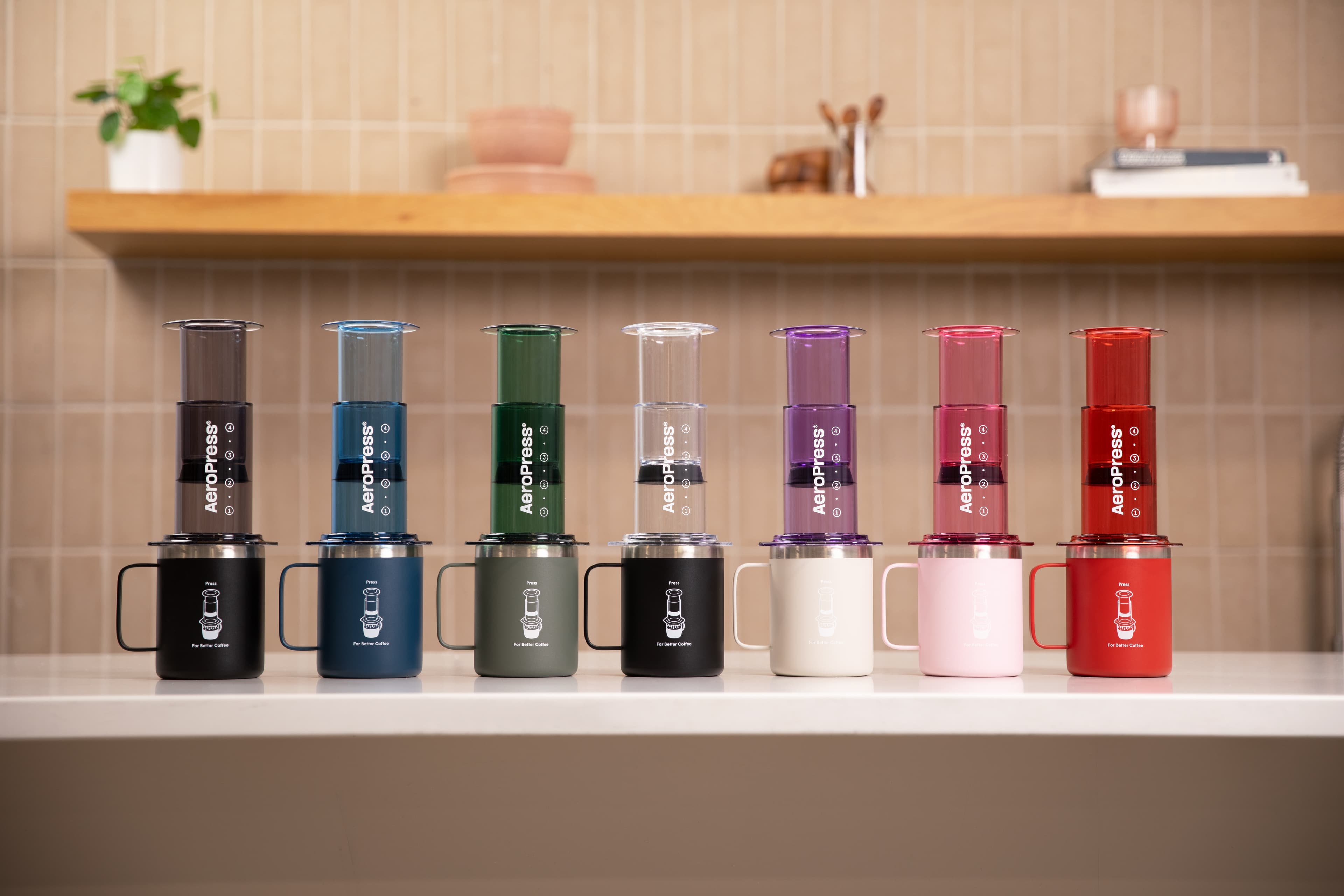 Siete cafeteras AeroPress de colores vibrantes alineadas sobre tazas a juego en un mostrador blanco.