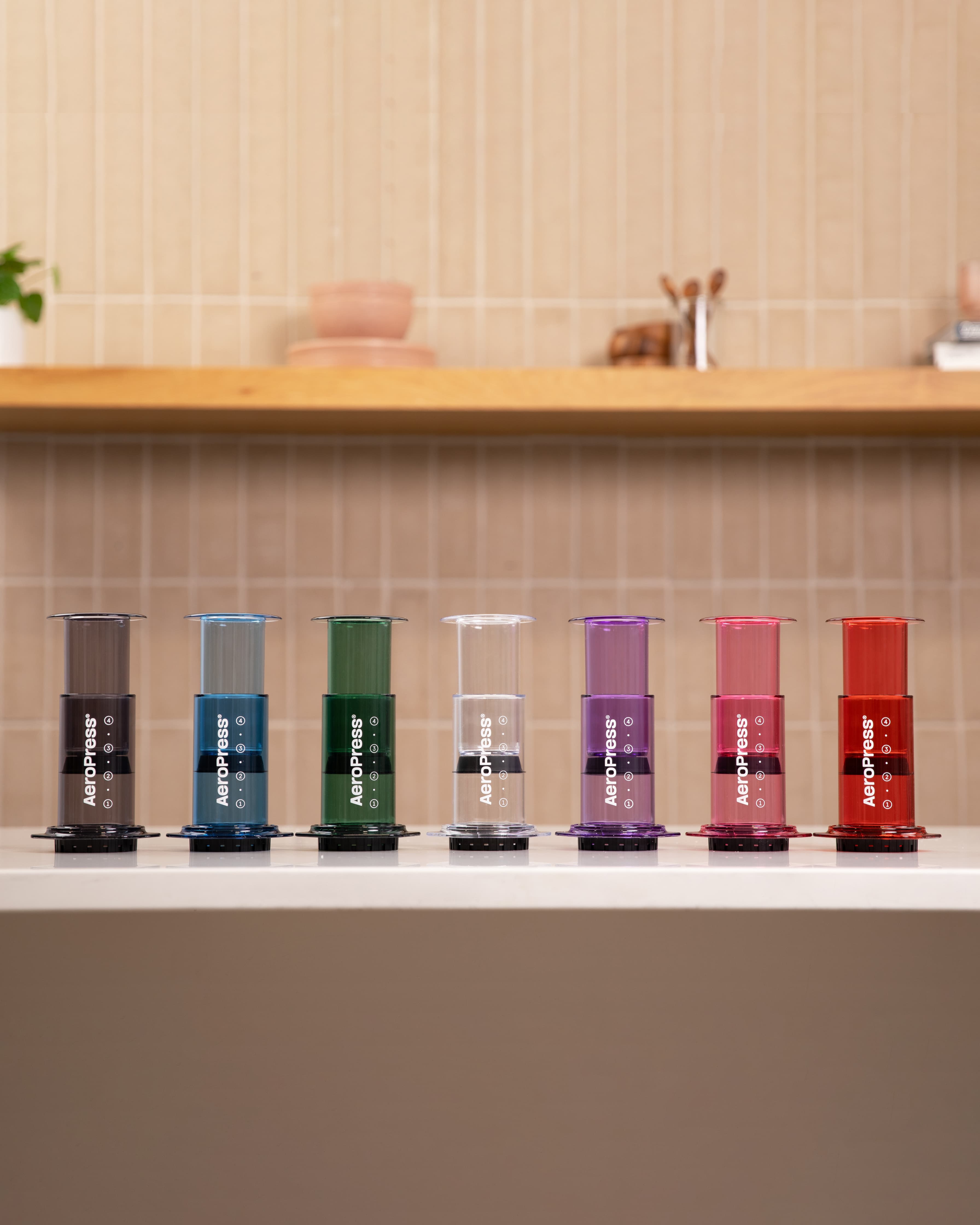 Siete cafeteras AeroPress de distintos colores transparentes alineadas sobre una encimera blanca de cocina.