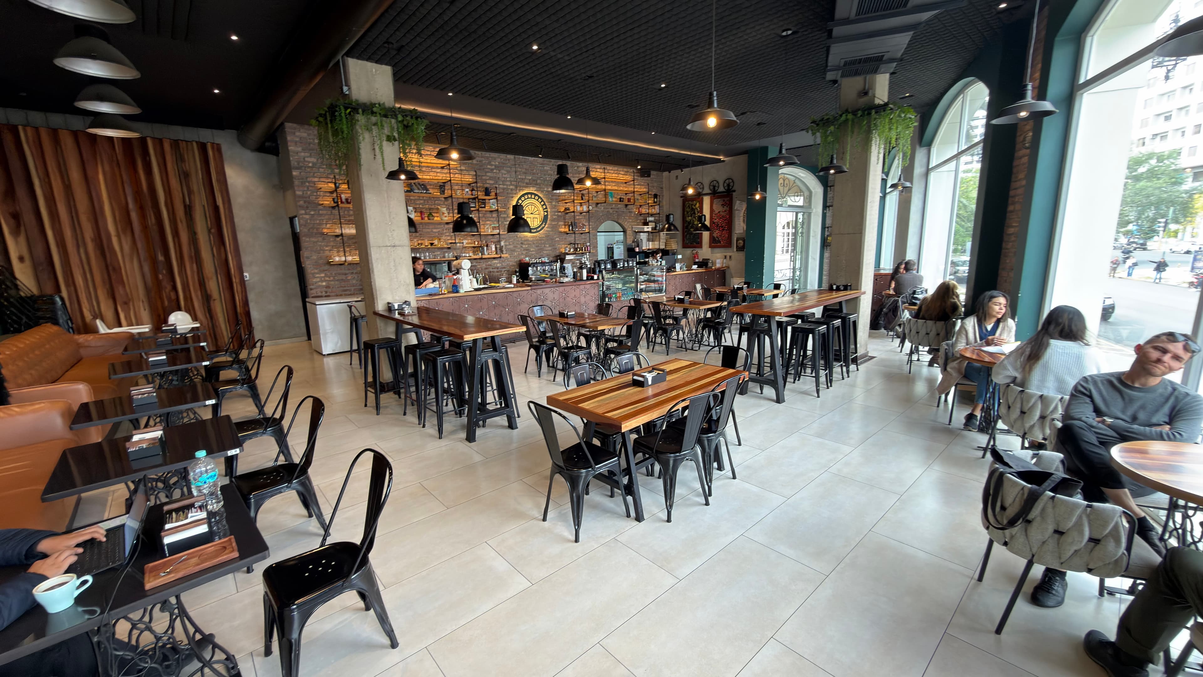 Cafetería industrial con paredes de ladrillo, mesas de madera, sillas negras y grandes ventanales luminosos.