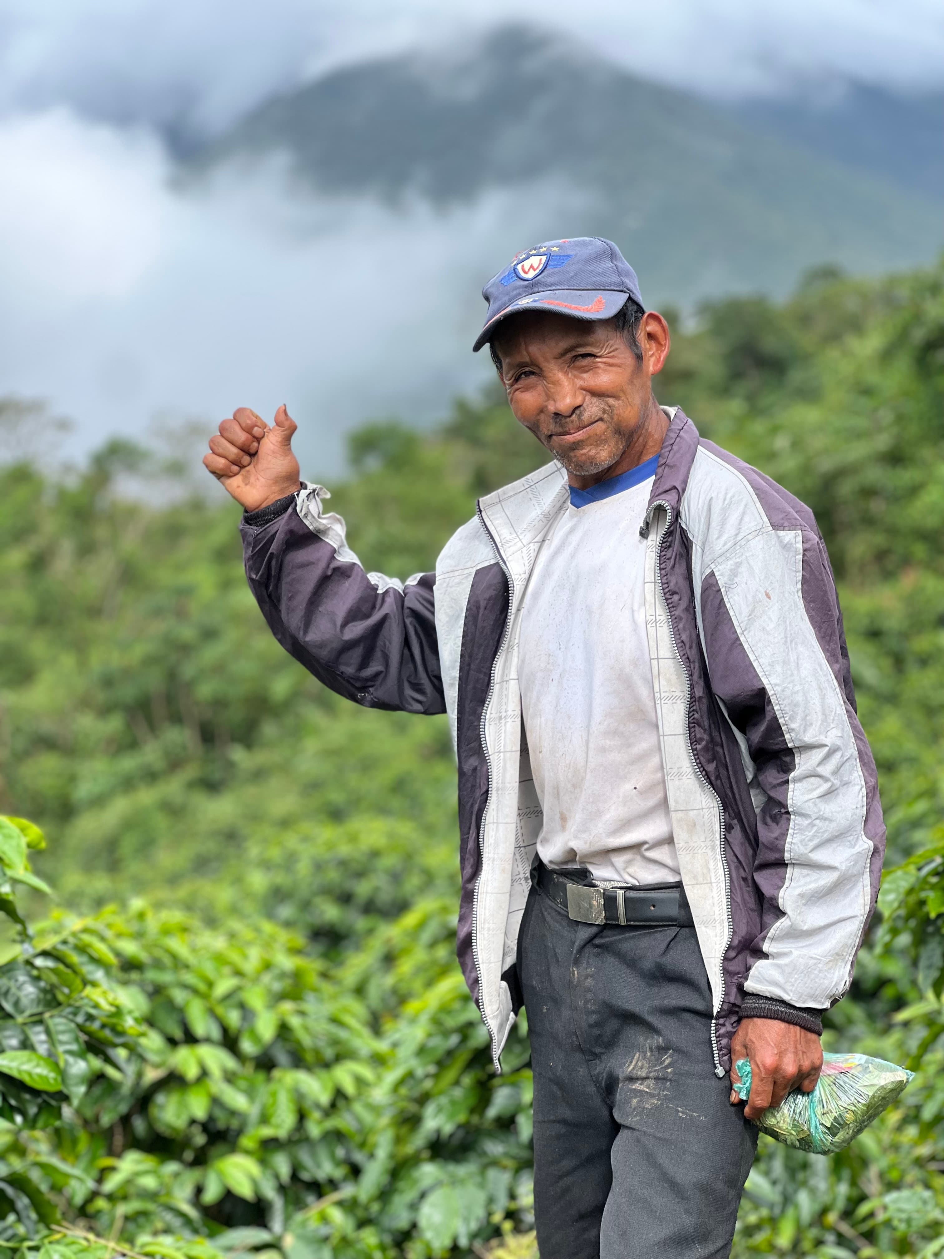 Campesino sonriente alzando el puño en un cafetal verde con montañas brumosas de fondo.