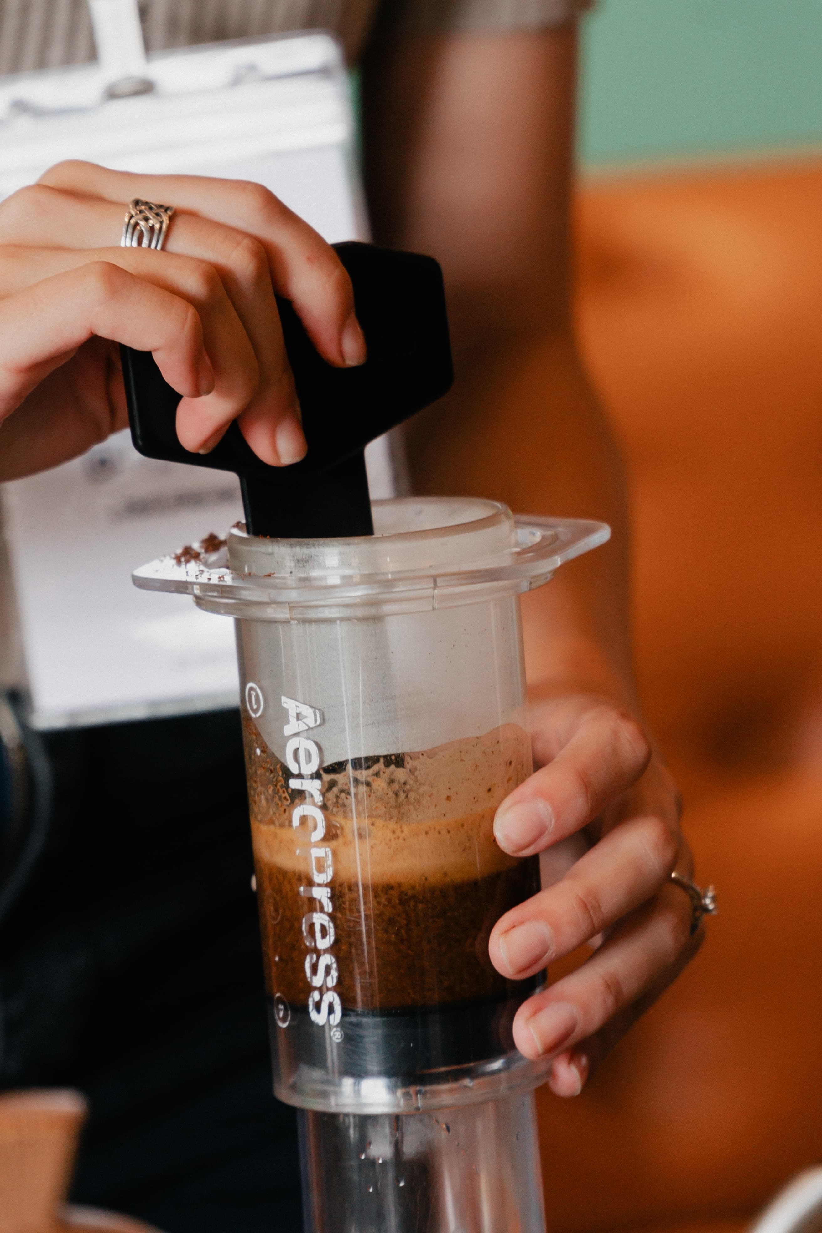 Manos removiendo café molido con una paleta negra dentro de una cafetera AeroPress transparente.