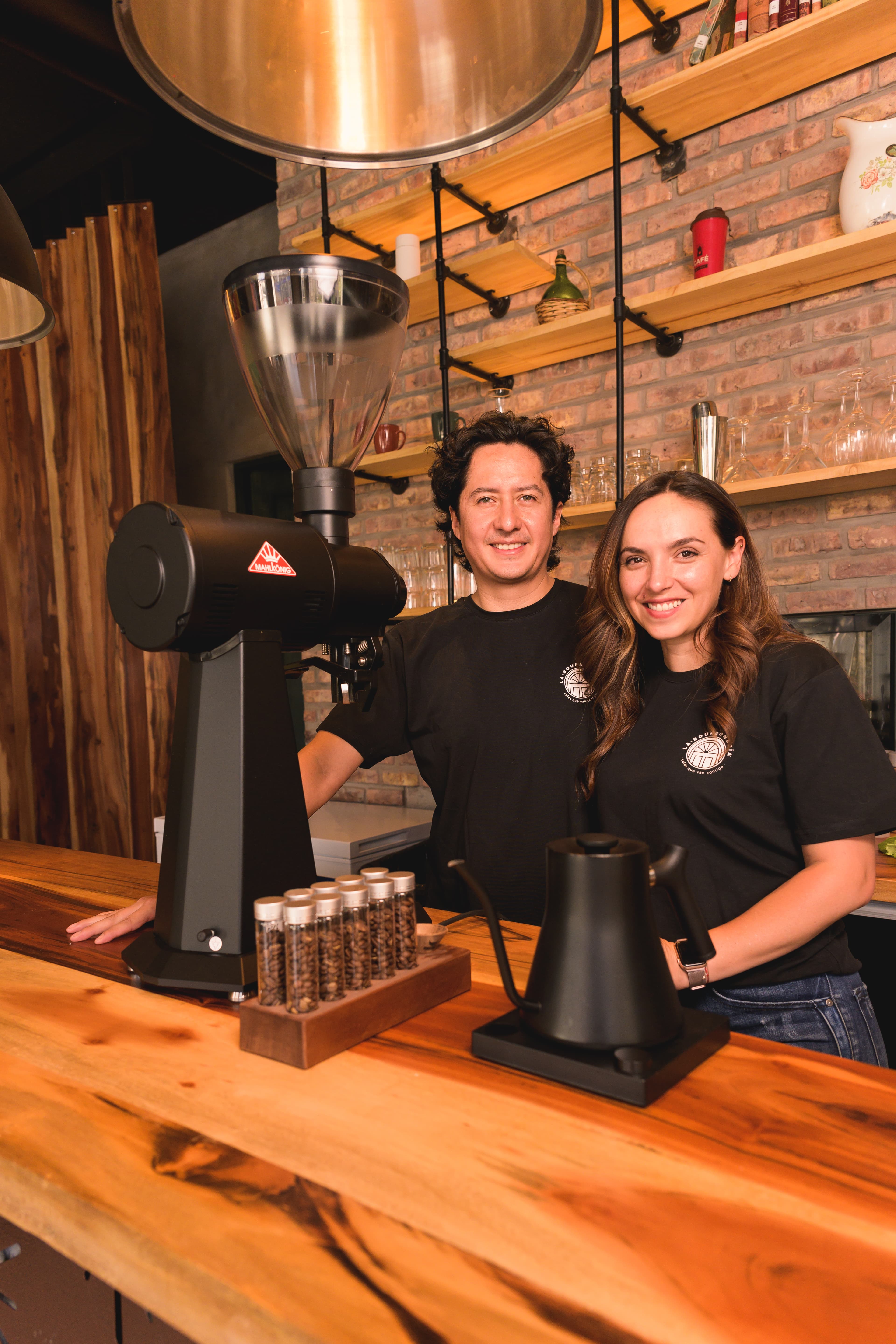 Pareja sonriente tras barra de madera con molinillo profesional y granos de café.