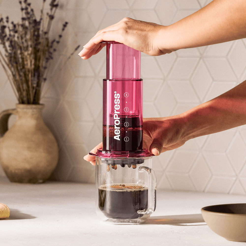 Manos presionando una cafetera AeroPress rosa para filtrar café en una taza de cristal transparente.