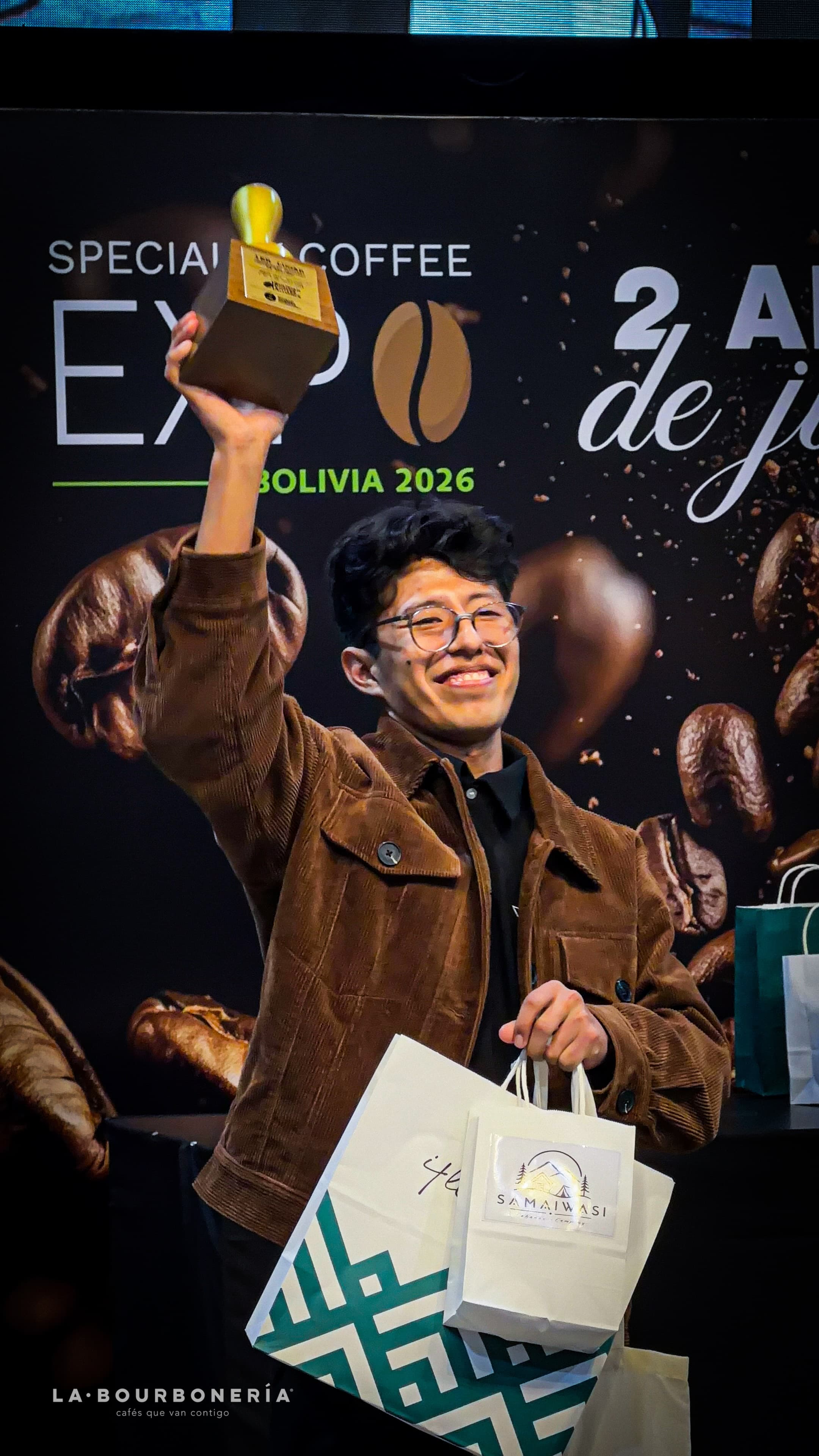Joven sonriente con chaqueta marrón alzando un trofeo en una feria de café especial.